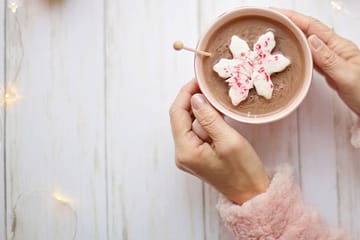 Cozy winter scene featuring a pair of hands in soft pink fluffy sleeves holding a pastel pink cup of hot chocolate. The drink is topped with a snowflake-shaped marshmallow sprinkled with crushed peppermint. A delicate wooden stirrer rests on the edge of the cup. The background consists of a white wooden surface decorated with warm fairy lights, adding a festive and comforting ambiance.