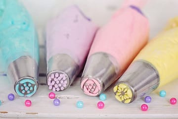 A close-up of four piping bags filled with pastel-colored frosting in shades of blue, purple, pink, and yellow, arranged on a rustic white wooden surface. Each bag has a decorative metal piping tip designed for creating different icing patterns. Small, round, glossy sprinkles in pink, blue, and purple are scattered around, adding a playful touch to the scene.