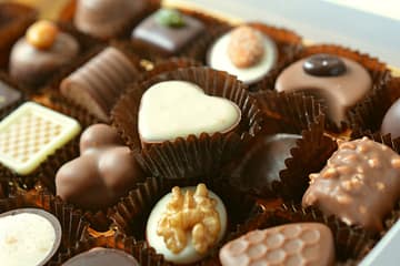 A close-up of an assorted box of gourmet chocolates, each placed in a brown paper cup. The selection includes a white chocolate heart, a dark chocolate with a walnut topping, and various milk and dark chocolate pieces with intricate designs, nuts, and decorative patterns. The chocolates are arranged neatly in a luxurious gift box.
