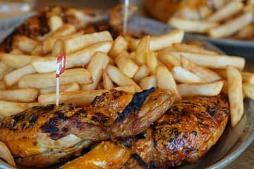 Close-up of a plate filled with golden, crispy French fries and grilled chicken with a spicy, glazed coating. The chicken has a slightly charred surface and is marked with a small red flag indicating a hot spice level. The dish is served on a rustic wooden table, with another plate of similar food slightly blurred in the background.