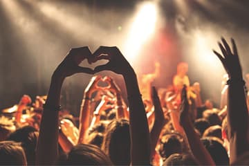 Vibrant concert scene with a crowd raising their hands in the air. A person in the foreground forms a heart shape with their hands, while others in the audience do the same. The stage is illuminated with warm, golden lights, and a performer is seen singing in the background, slightly out of focus.